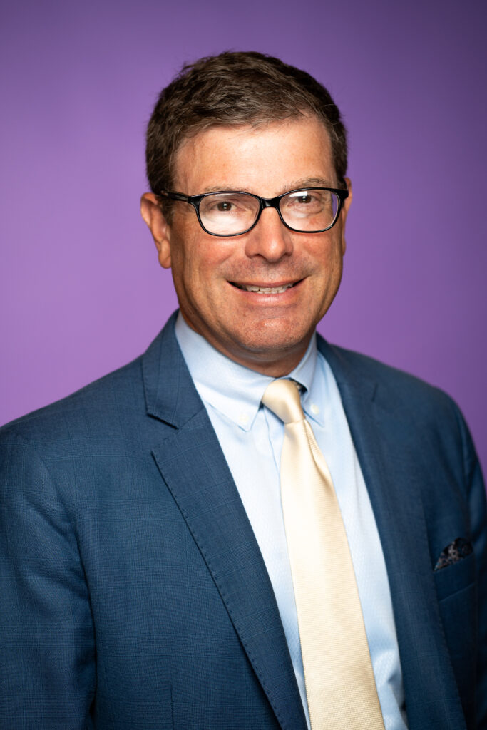 John Meko - A man in a suit with glasses in front of a purple background